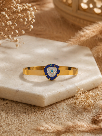 Antica Designs flat gold-toned cuff bracelet featuring a shield-shaped centerpiece with azure blue enamel, crystal pavé, and an evil eye motif displayed on a hexagonal stone plinth.
