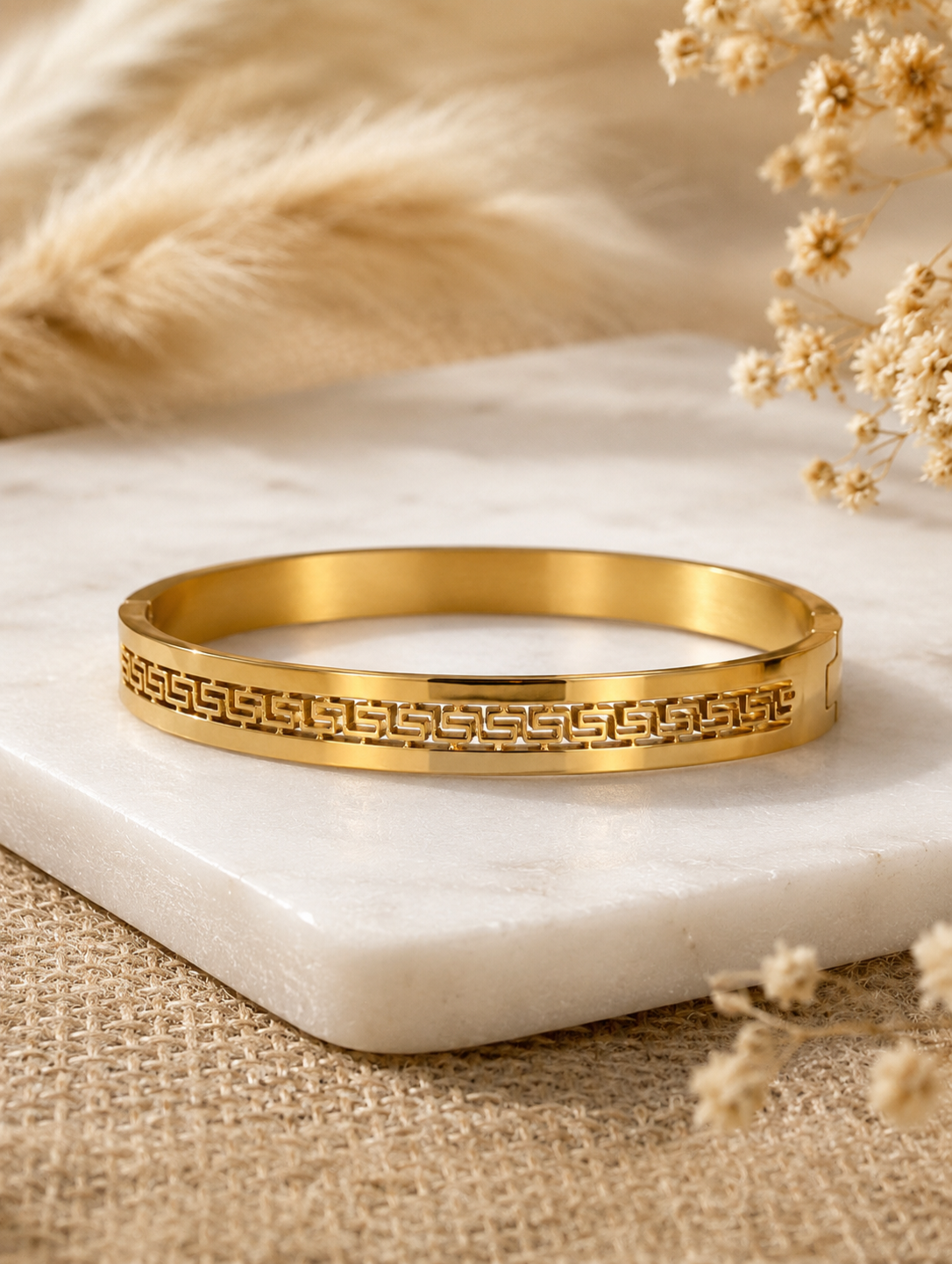 Antica Designs premium high-polish gold-toned hinged bangle featuring a central cutout Greek key geometric pattern, displayed elegantly on a square white marble slab.