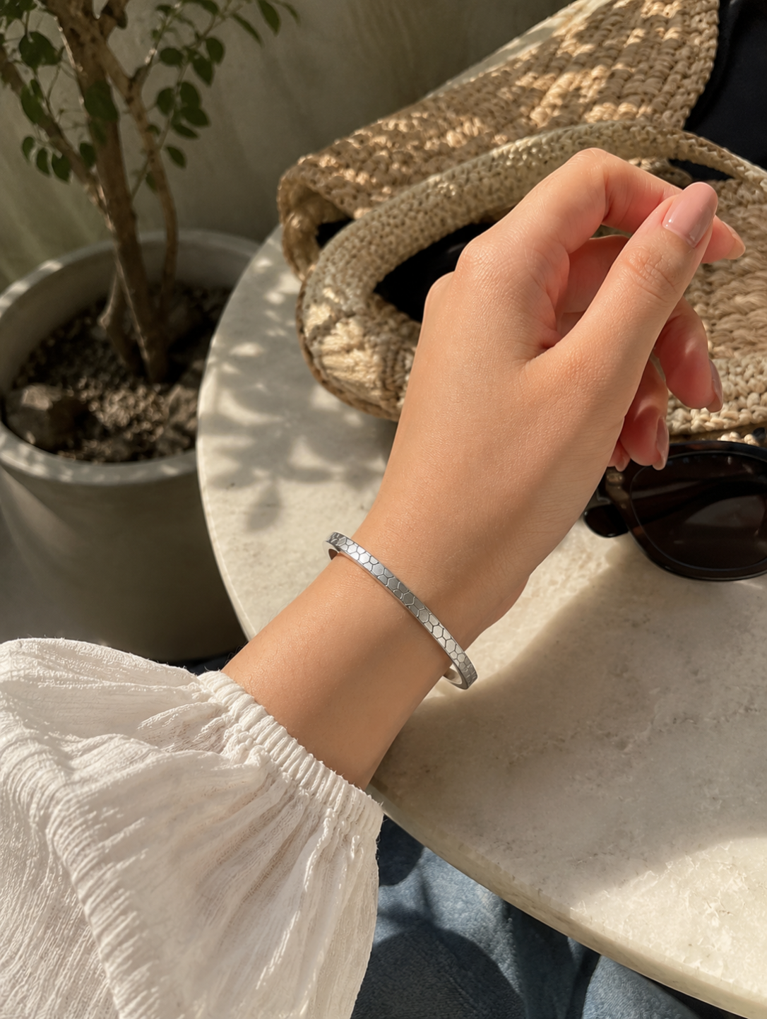 Antica Designs sleek, silver-toned thin bangle with an engraved geometric honeycomb texture, worn on a wrist in a bright, modern outdoor cafe setting.