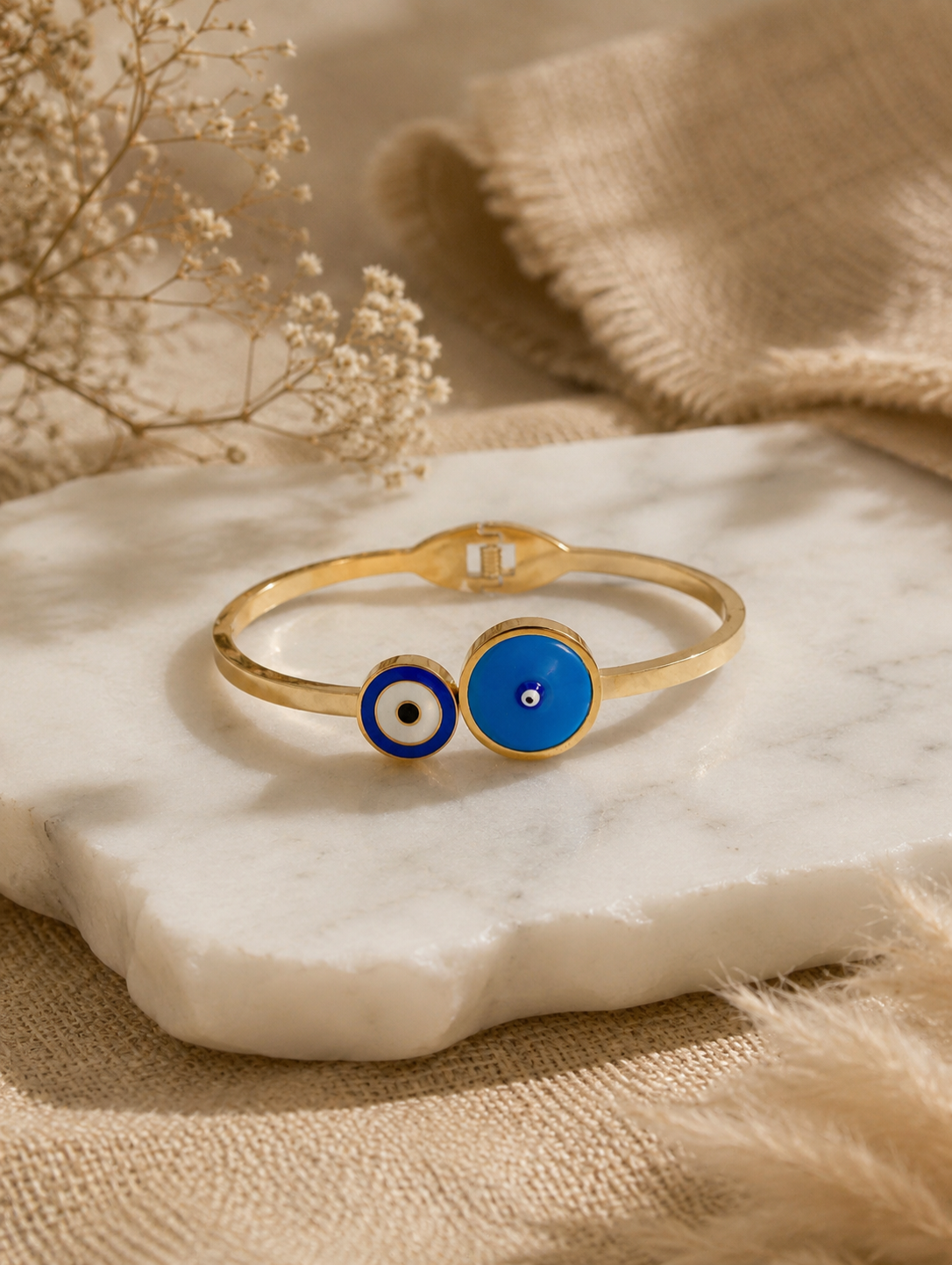 Antica Designs gold-toned hinged bangle featuring an asymmetric dual-medallion evil eye design in sky blue and dark blue enamel, styled on a white marble surface.