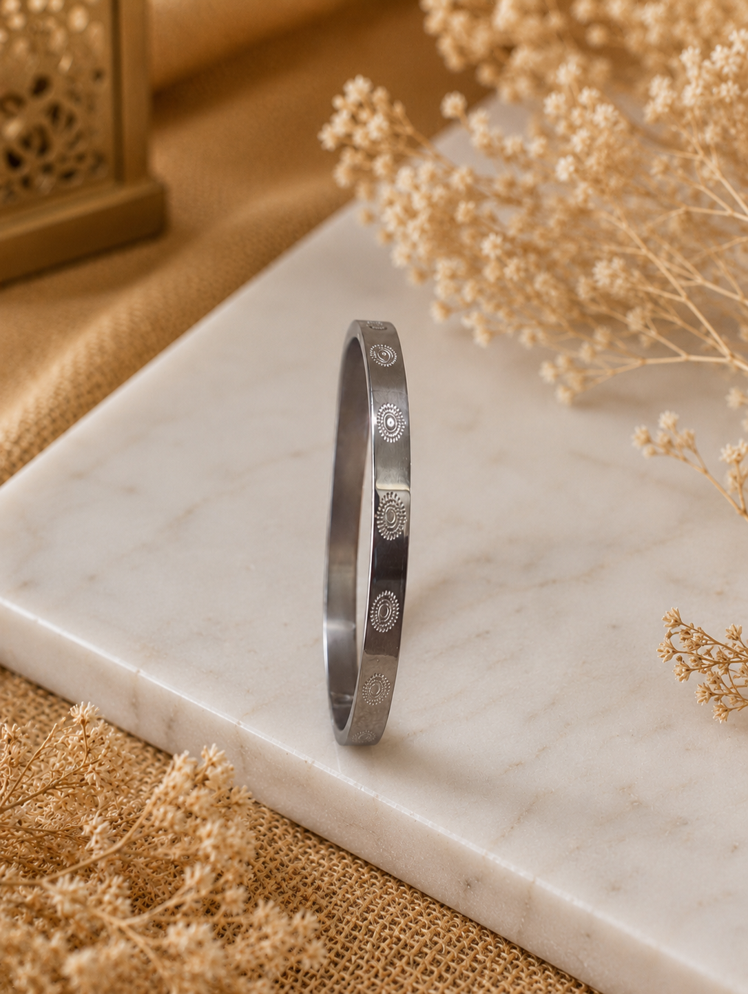 Antica Designs sleek silver-toned flat band bangle with a repeating engraved sunburst pattern, displayed upright on a white marble slab