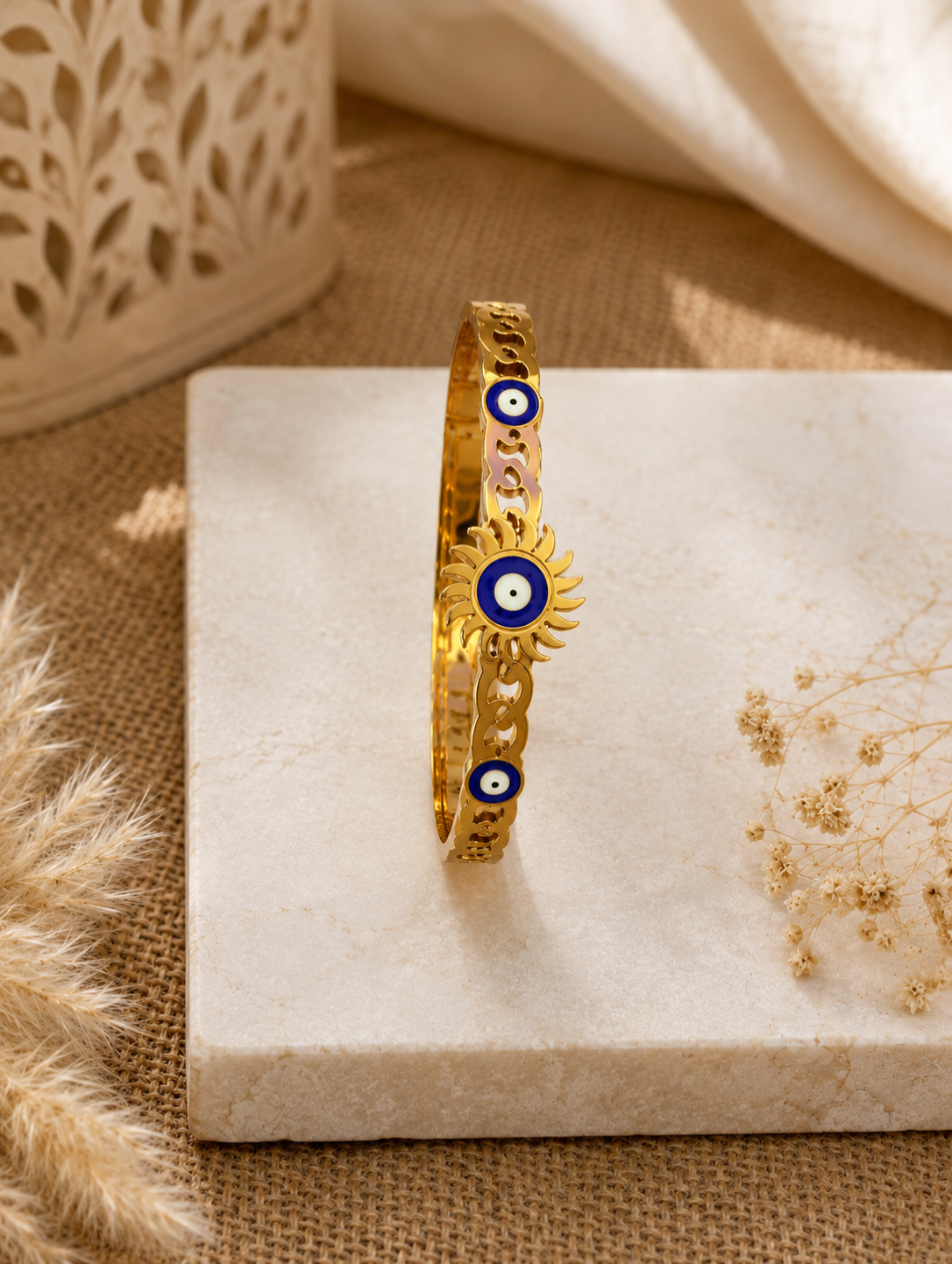 Antica Designs gold-toned hinged bangle featuring an open-work chain band and an enamel sunburst evil eye centerpiece, displayed on a square white marble slab.