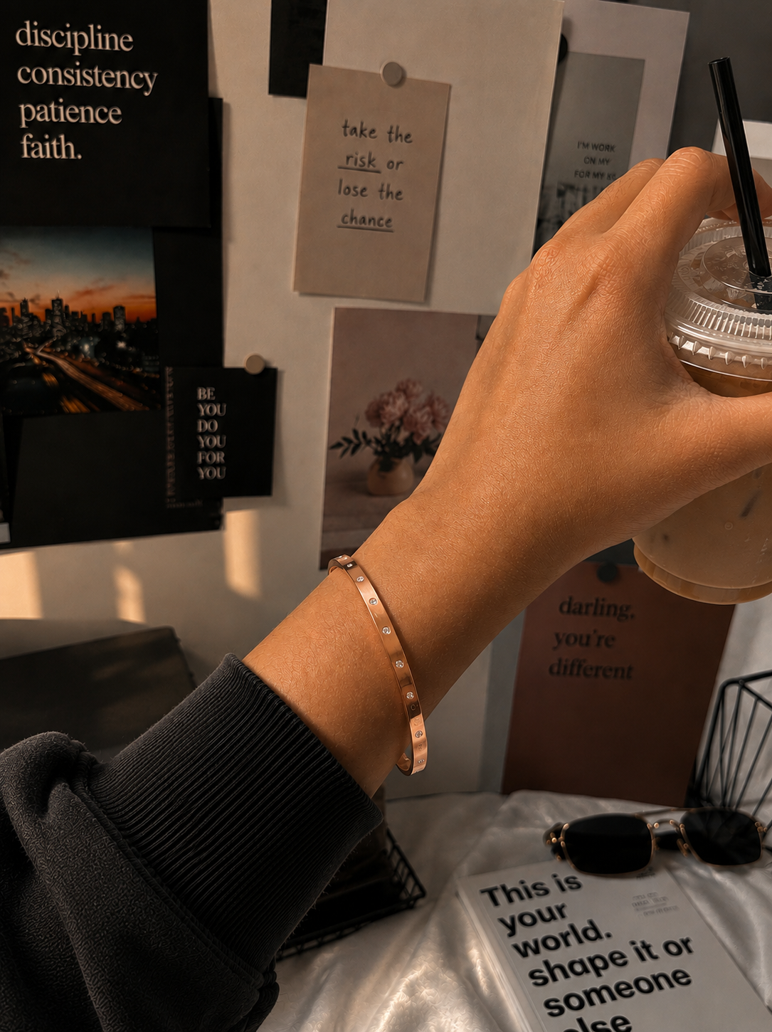 A lifestyle close-up shot for the Gemdot collection, featuring a person's wrist adorned with a sleek rose-gold bangle bracelet embedded with small, sparkling crystals. The hand is holding an iced coffee cup against an aesthetic background of motivational wall prints, sunglasses, and a book. As seen in KD022_gemdot_2.png.