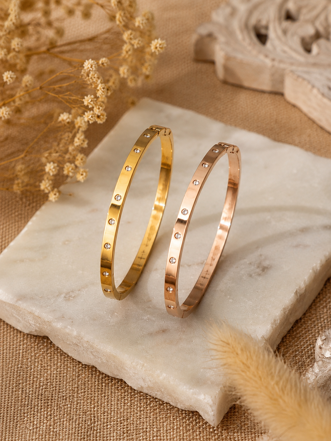 A lifestyle close-up shot for the Gemdot collection, featuring a person's wrist adorned with a sleek rose-gold bangle bracelet embedded with small, sparkling crystals. The hand is holding an iced coffee cup against an aesthetic background of motivational wall prints, sunglasses, and a book. As seen in KD022_gemdot_2.png