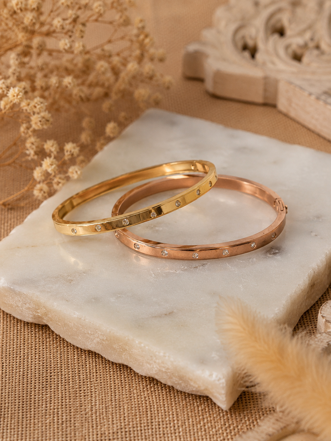 A lifestyle close-up shot for the Gemdot collection, featuring a person's wrist adorned with a sleek rose-gold bangle bracelet embedded with small, sparkling crystals. The hand is holding an iced coffee cup against an aesthetic background of motivational wall prints, sunglasses, and a book. As seen in KD022_gemdot_2.png