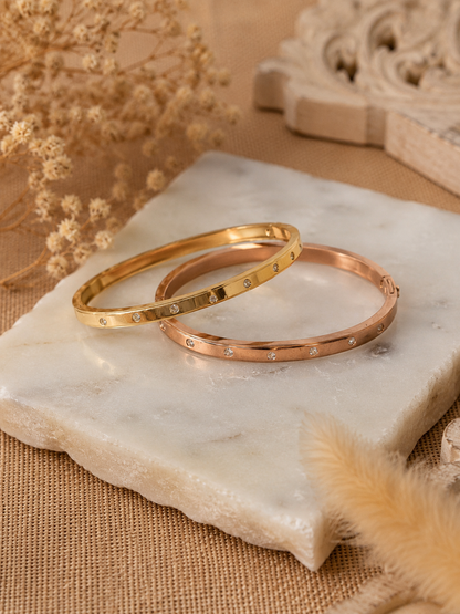 A lifestyle close-up shot for the Gemdot collection, featuring a person's wrist adorned with a sleek rose-gold bangle bracelet embedded with small, sparkling crystals. The hand is holding an iced coffee cup against an aesthetic background of motivational wall prints, sunglasses, and a book. As seen in KD022_gemdot_2.png