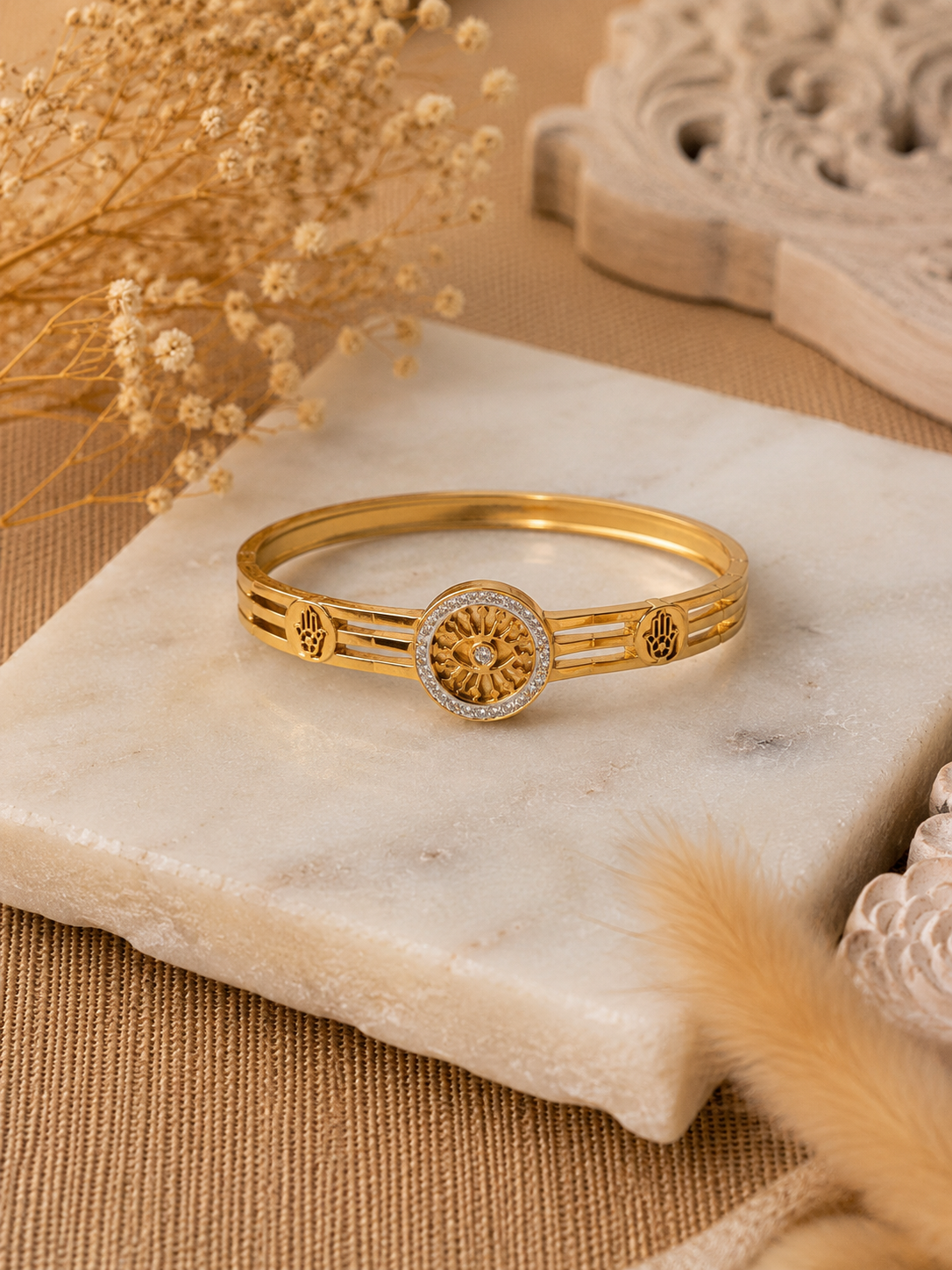 A flat-lay product photograph of a polished yellow-gold bangle bracelet resting on a textured white marble slab. The bangle features a central, circular sunwheel design with an evil eye center, bordered by small sparkling crystals, flanked by open-work Hamsa hand motifs on a triple-band frame. The background includes dried pampas grass and neutral woven textures. As seen in KD0023_sunwheel_a.png