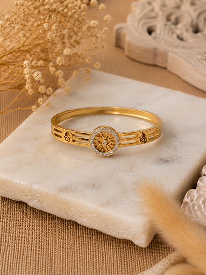 A flat-lay product photograph of a polished yellow-gold bangle bracelet resting on a textured white marble slab. The bangle features a central, circular sunwheel design with an evil eye center, bordered by small sparkling crystals, flanked by open-work Hamsa hand motifs on a triple-band frame. The background includes dried pampas grass and neutral woven textures. As seen in KD0023_sunwheel_a.png