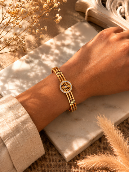 A flat-lay product photograph of a polished yellow-gold bangle bracelet resting on a textured white marble slab. The bangle features a central, circular sunwheel design with an evil eye center, bordered by small sparkling crystals, flanked by open-work Hamsa hand motifs on a triple-band frame. The background includes dried pampas grass and neutral woven textures. As seen in KD0023_sunwheel_a.png