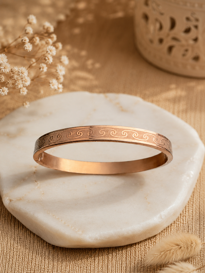 A flat-lay product photograph of a sleek rose-gold kada bracelet resting flat on a square white marble slab. The rigid bangle features an elegantly engraved continuous ocean wave and scroll motif along its outer band, framed by polished edges. The background is styled with dried baby's breath flowers, neutral tan woven textiles, and a carved wooden accent. As seen in KD024_rose gold kada_a.png.