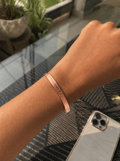 A flat-lay product photograph of a sleek rose-gold kada bracelet resting flat on a square white marble slab. The rigid bangle features an elegantly engraved continuous ocean wave and scroll motif along its outer band, framed by polished edges. The background is styled with dried baby's breath flowers, neutral tan woven textiles, and a carved wooden accent. As seen in KD024_rose gold kada_a.png.