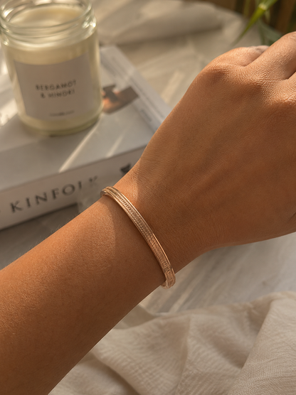 A lifestyle close-up photograph for the Doro collection, showing a person's wrist wearing a slender, textured gold-toned bangle bracelet bathed in warm sunlight. The background features a cozy setup with a Kinfolk book, a jar candle labeled "Bergamot & Mimosa", and soft linen fabrics. As seen in KD025_doro.png.