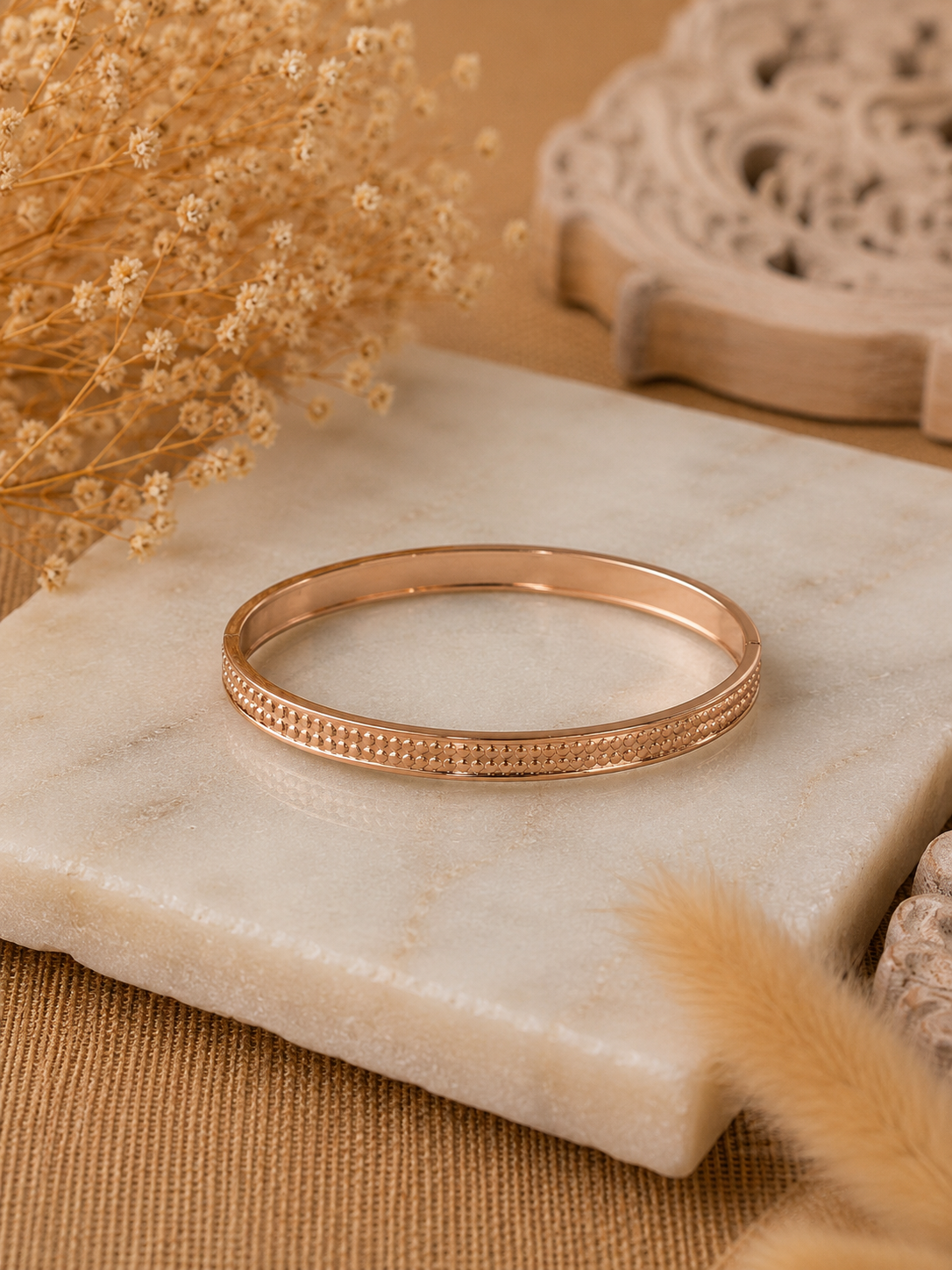 A lifestyle close-up photograph for the Doro collection, showing a person's wrist wearing a slender, textured gold-toned bangle bracelet bathed in warm sunlight. The background features a cozy setup with a Kinfolk book, a jar candle labeled "Bergamot & Mimosa", and soft linen fabrics. As seen in KD025_doro.png.