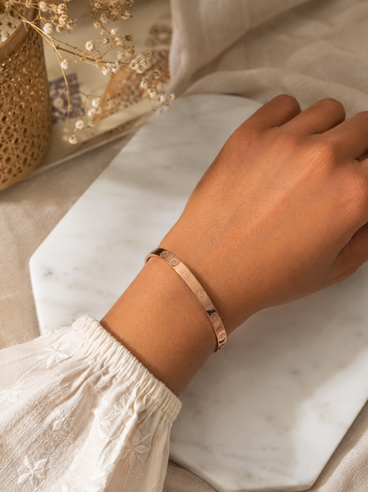 A product photograph of the Rosario rose gold kada bracelet standing vertically on an angle over a square white marble block. The rigid bangle showcases evenly spaced, engraved circular medallion designs on its polished flat surface. The background is styled with warm neutral tones, featuring dried pampas grass, delicate baby's breath flowers, and soft burlap textures. As seen in KD026_minimal design kada_350.png.