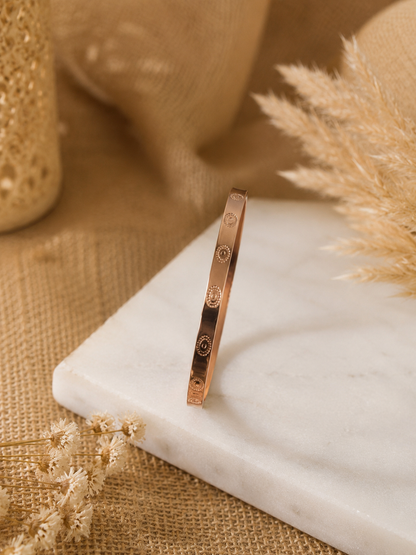 A product photograph of the Rosario rose gold kada bracelet standing vertically on an angle over a square white marble block. The rigid bangle showcases evenly spaced, engraved circular medallion designs on its polished flat surface. The background is styled with warm neutral tones, featuring dried pampas grass, delicate baby's breath flowers, and soft burlap textures. As seen in KD026_minimal design kada_350.png.