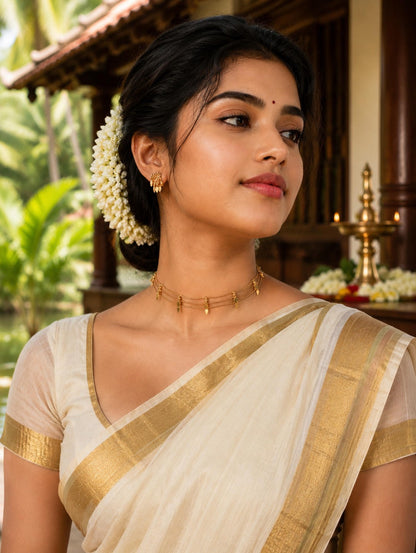 Woman in a traditional white saree with gold border, wearing gold jewelry, in a tropical setting.