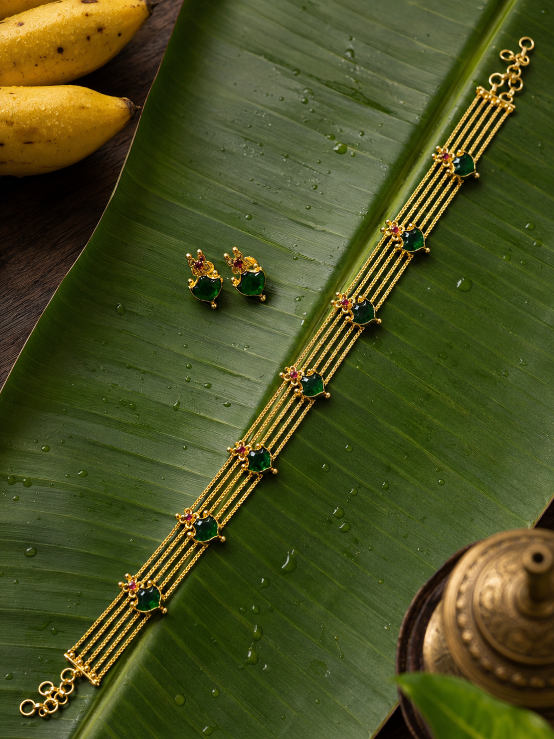 A traditional South Indian style gold jewellery set including a four-strand choker necklace and small matching stud earrings, both adorned with vibrant heart-shaped green stones and tiny red accents, displayed on a wet green banana leaf.