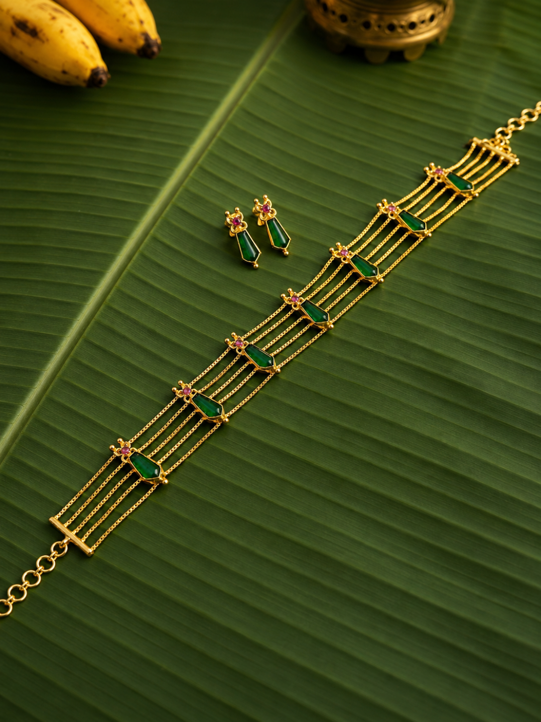 A traditional gold jewellery set named Vaidehi, featuring a four-strand choker necklace and matching drop earrings adorned with elongated, geometric green stones and small red gemstone accents, arranged on a green banana leaf.