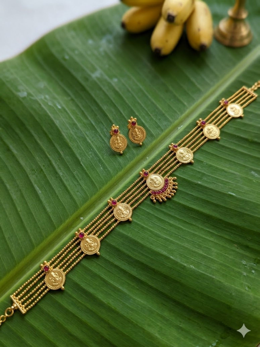 The Malavika traditional gold jewellery set featuring a multi-strand choker necklace adorned with circular Lakshmi coins, small pink stone accents, and a central semi-circular pendant, accompanied by matching round coin stud earrings on a green banana leaf.