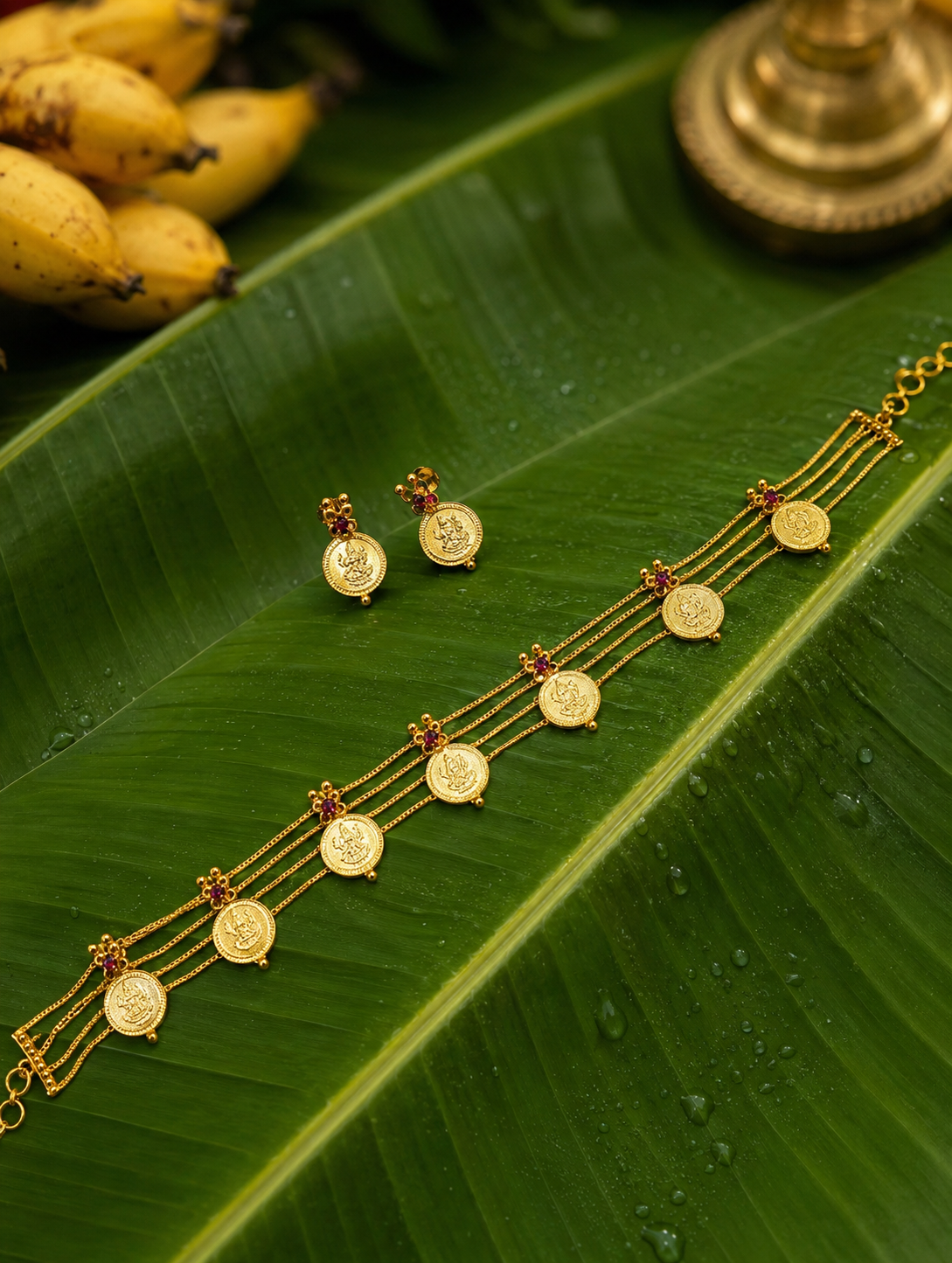 A traditional South Indian gold jewelry set, featuring a three-layered Kasu Mala choker necklace and matching stud earrings adorned with Goddess Lakshmi coins and small red ruby gemstones, displayed on a fresh green banana leaf with water droplets.