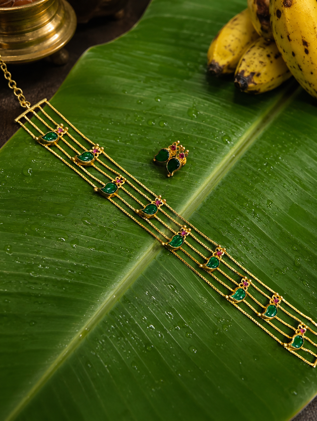 A traditional South Indian gold jewelry set from the Nandana collection, featuring a four-layered gold choker necklace and matching stud earrings decorated with green mango-shaped enamel motifs and tiny pinkish-red gemstones, laid flat on a wet green banana leaf.