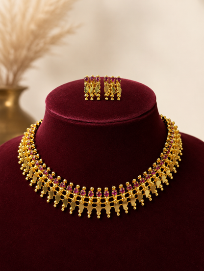 A traditional South Indian antique gold jewelry set displayed on a dark maroon velvet bust, featuring a collar-style gold choker necklace embedded with a row of pinkish-red ruby gemstones and hanging gold leaf drops, with matching square-shaped ruby stud earrings placed on top.