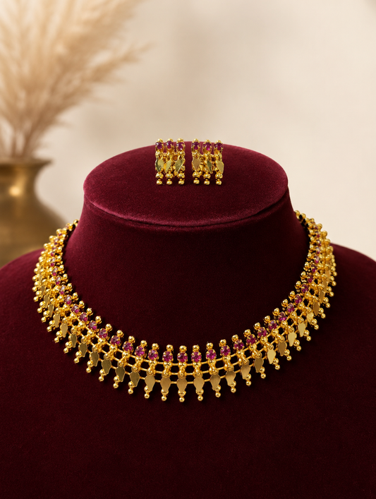 A traditional South Indian antique gold jewelry set displayed on a dark maroon velvet bust, featuring a collar-style gold choker necklace embedded with a row of pinkish-red ruby gemstones and hanging gold leaf drops, with matching square-shaped ruby stud earrings placed on top.