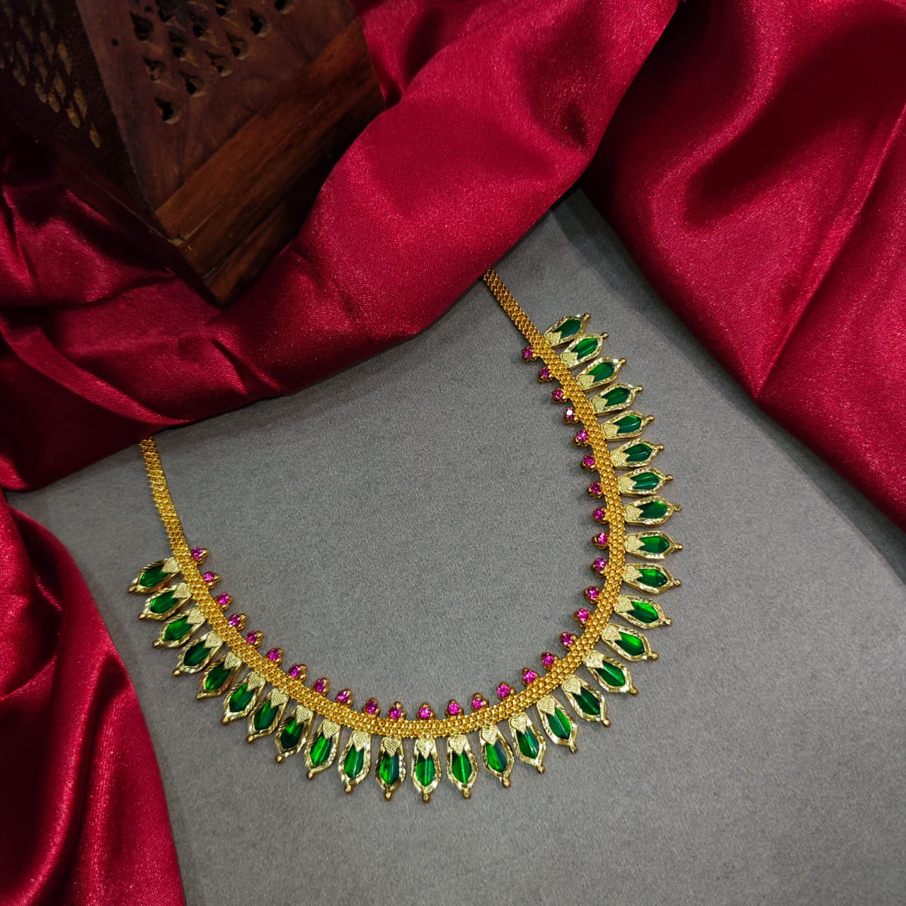 Gold necklace with green and red stones on a gray surface with red fabric