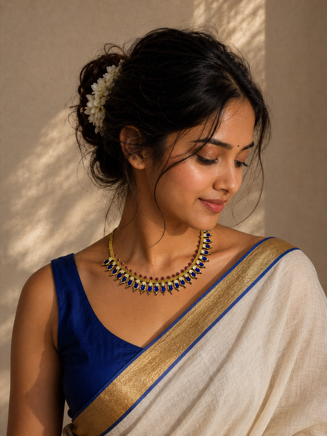 A close-up of a model wearing the Prajwala royal blue and cream enamel gold choker necklace. She is styled in a cream handloom saree with a blue blouse and a flower-adorned bun under warm, dappled sunlight.