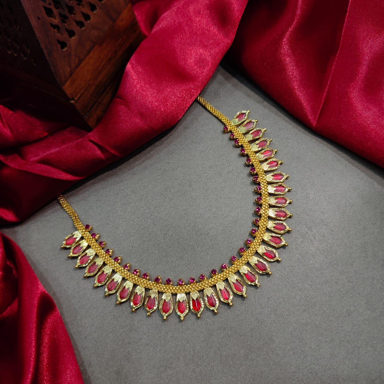 Traditional South Indian gold choker necklace from the Prajwala collection, featuring pointed ruby-red stone charms and small red accents, displayed flat on a grey background bordered by a rich red silk fabric.