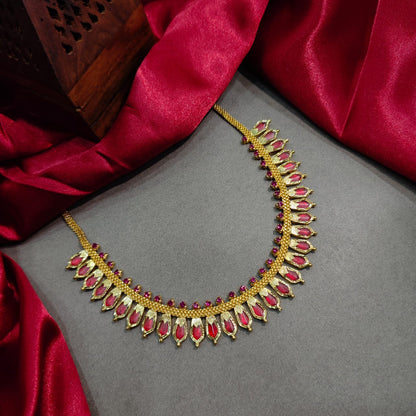Traditional South Indian gold choker necklace from the Prajwala collection, featuring pointed ruby-red stone charms and small red accents, displayed flat on a grey background bordered by a rich red silk fabric.