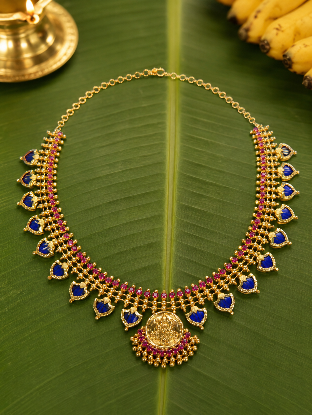 Royal blue stone variant of the traditional Kerala Palakka necklace laid flat on a banana leaf, highlighted by blue leaf-shaped stone and a central golden deity pendant.