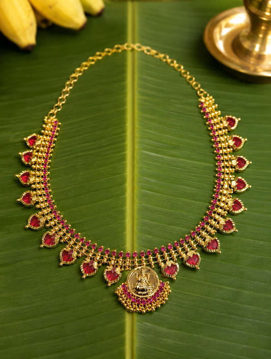 Traditional South Indian Palakka necklace flat lay on a green banana leaf, featuring green leaf-shaped stones, bordered by ruby pink accents and a central golden Lakshmi coin pendant.