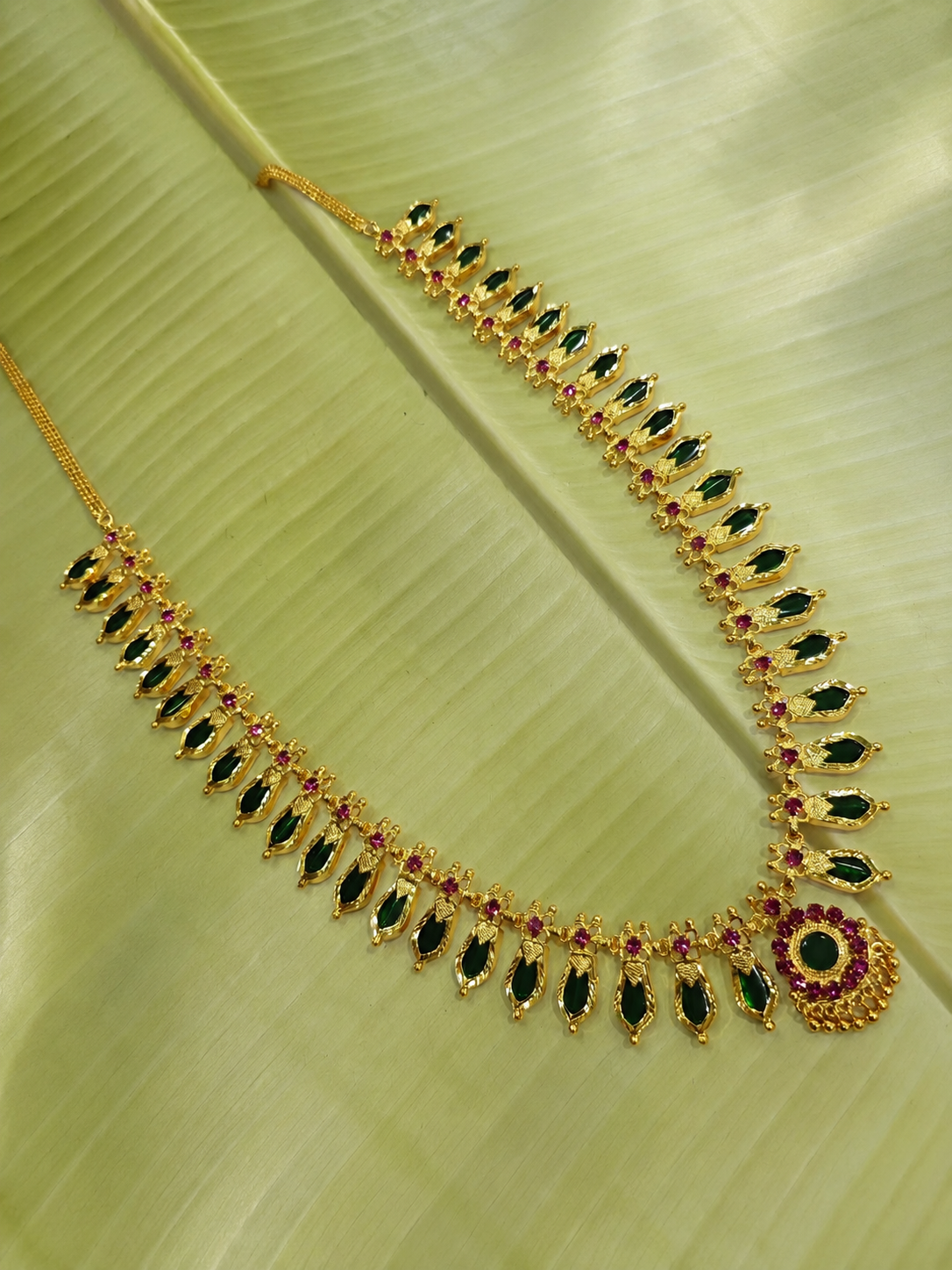Classic emerald green stone variant of the South Indian Nagapadam gold necklace arranged neatly on a light green banana leaf background.