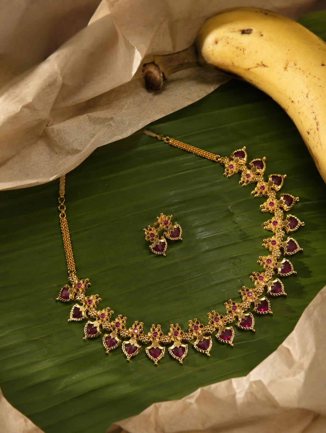 A ruby red leaf-shaped Kerala style Palakka necklace set with matching earrings, arranged on a banana leaf background with a yellow banana and decorative parchment paper.