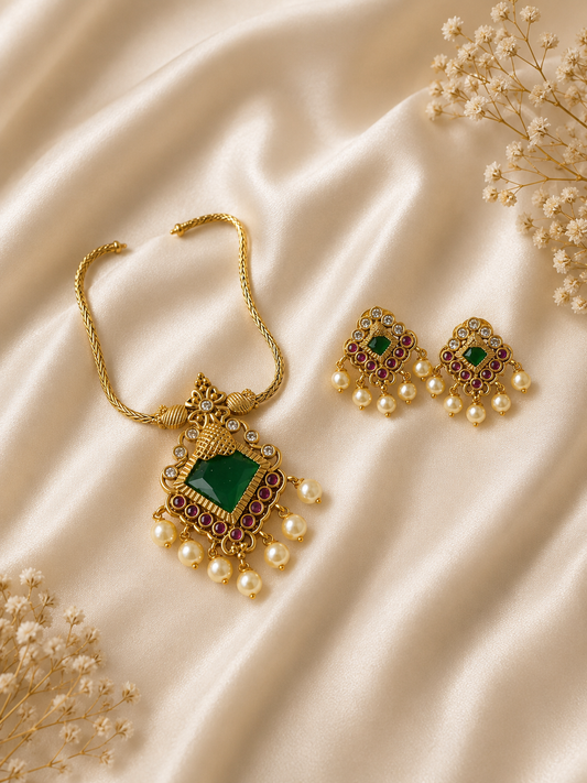 Flat lay view of an antique gold-plated jewelry set featuring a diamond-shaped emerald green stone pendant necklace and matching jhumka-style earrings with pink ruby accents and hanging white pearls, beautifully arranged on a shimmering champagne silk background with delicate dried white flowers.