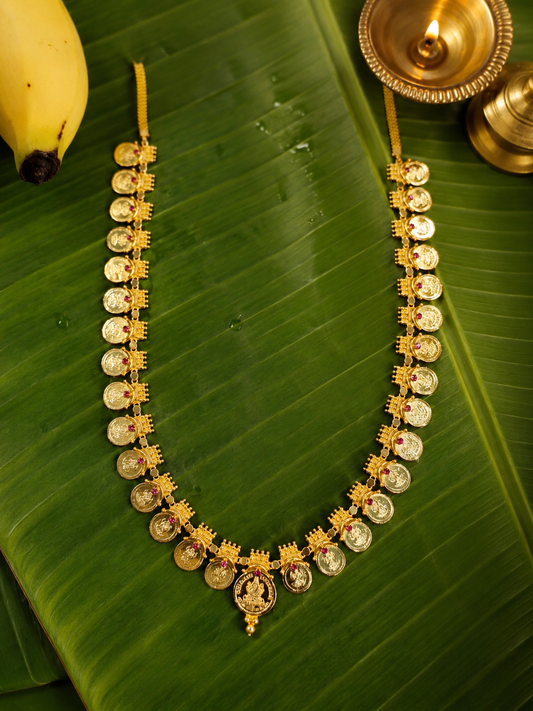 A grand bridal-style long Padmakshi Kasu Mala featuring thirty gold coins with a central Lakshmi pendant and ruby embellishments, arranged in a U-shape on a wet banana leaf with a traditional lit oil lamp.