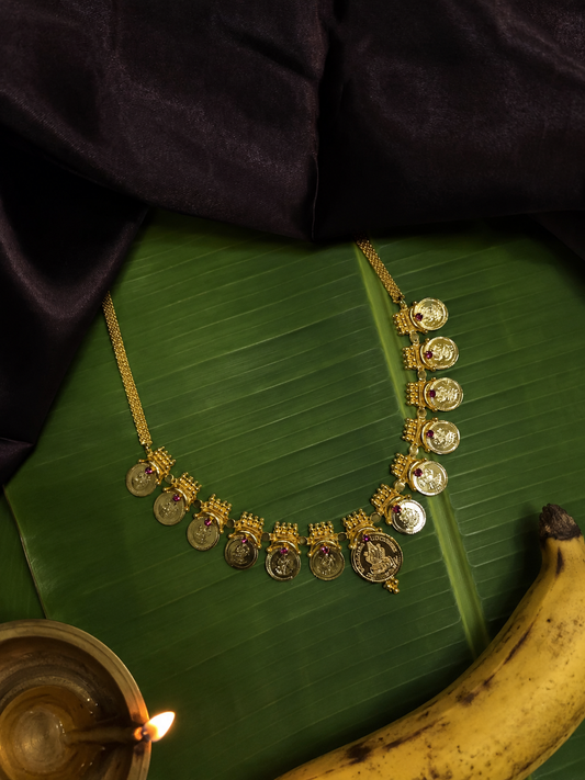 A classic South Indian Kasu Mala necklace adorned with twelve gold Lakshmi coins and ruby settings, displayed on a banana leaf alongside a brass oil lamp and a yellow banana beneath dark purple drapery.