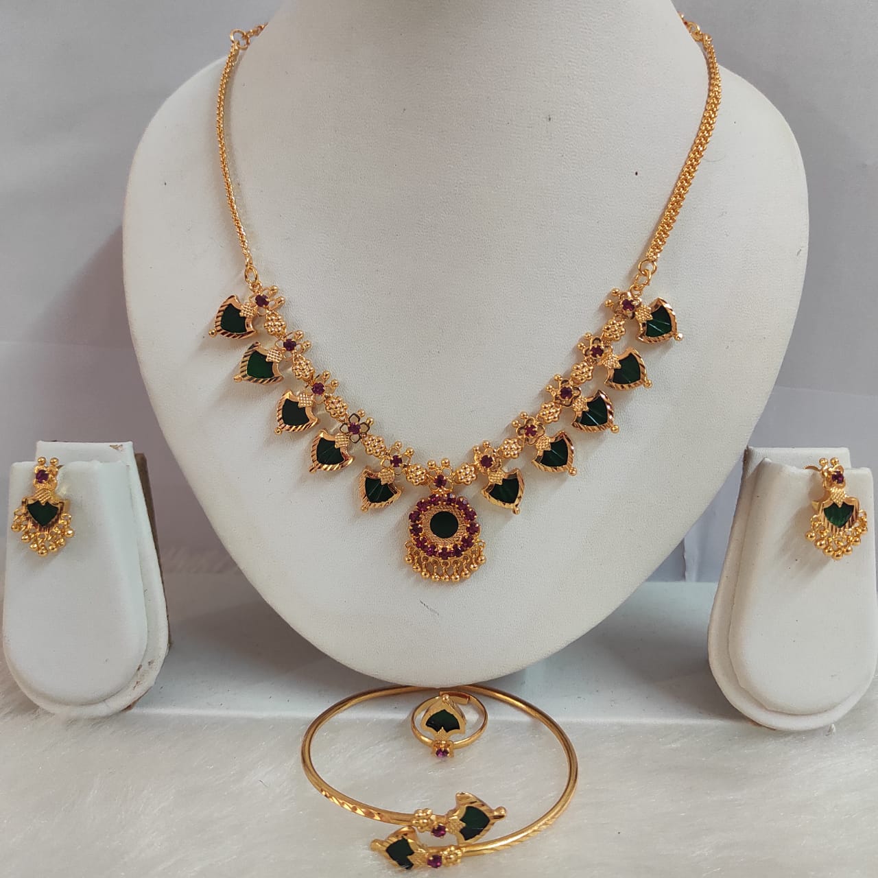 Gold necklace with green and red stones on a mannequin bust, accompanied by matching earrings and bangles.