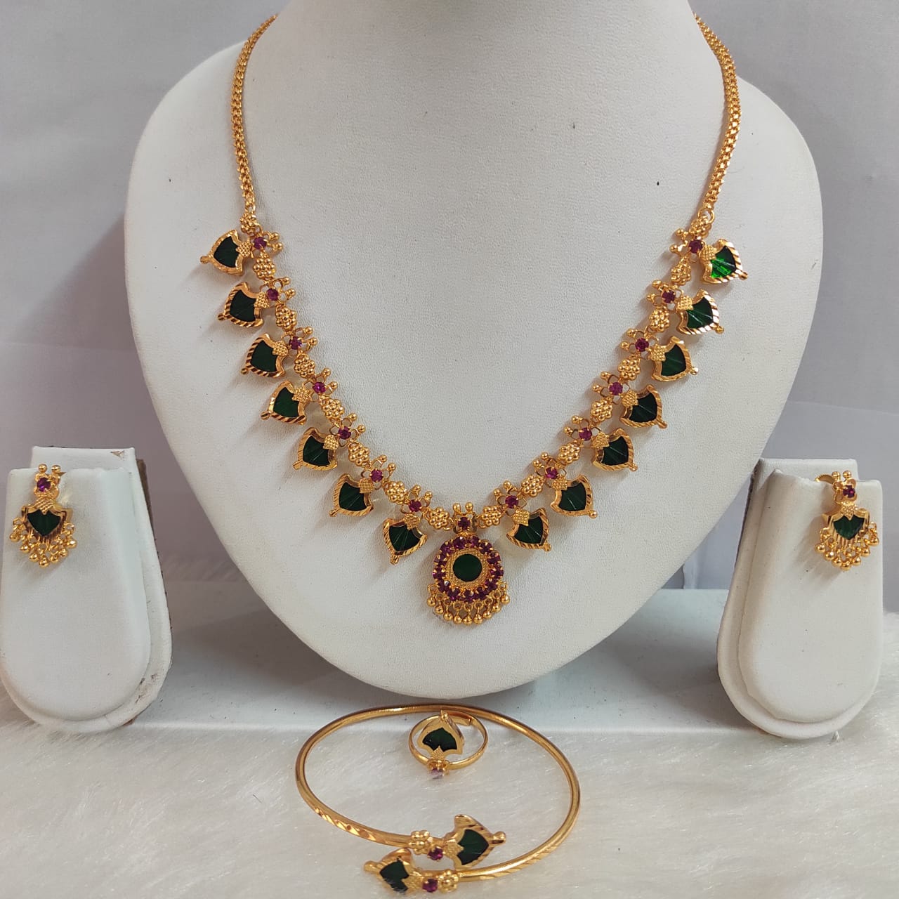 Gold necklace with green and red gemstones on a white stand, accompanied by earrings and a bracelet.