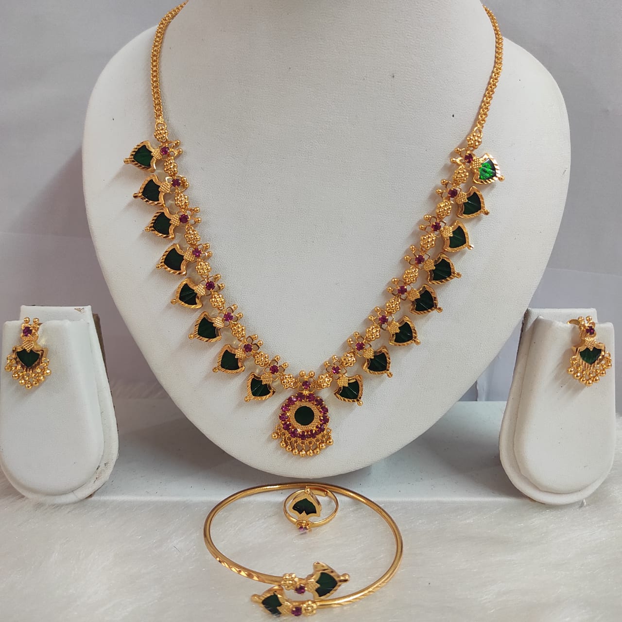 Gold necklace with green and red gemstones on a white stand, accompanied by matching earrings and bangles.