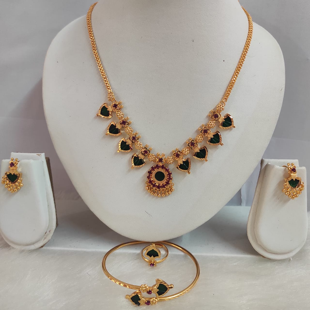 Gold necklace with green and red gemstones on a white stand, accompanied by matching earrings and bangles.