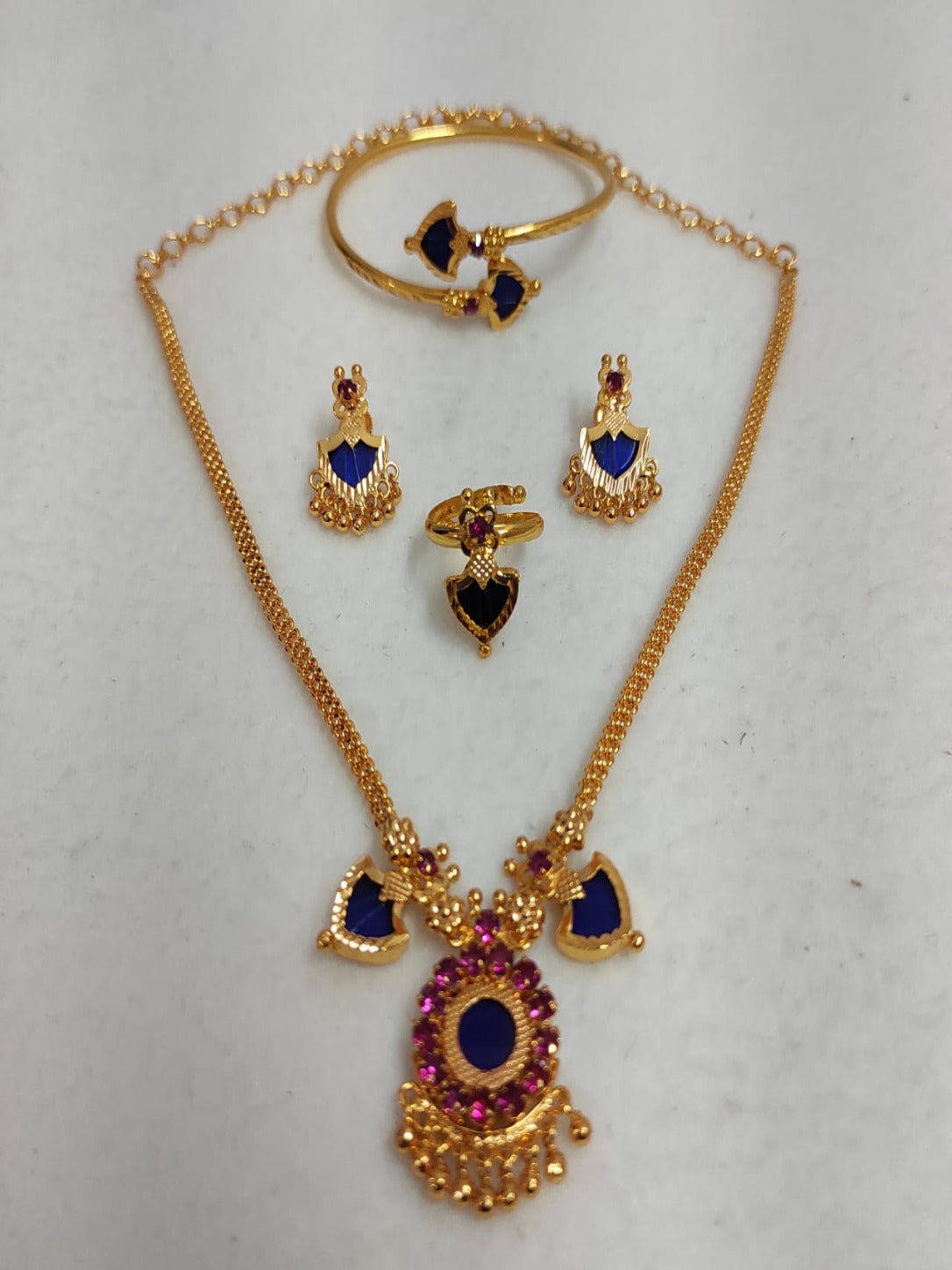 Nandika 2 Charm gold-plated jewelry set featuring a traditional necklace, matching earrings, a ring, and a bangle with deep sapphire blue and ruby stones on a white surface.