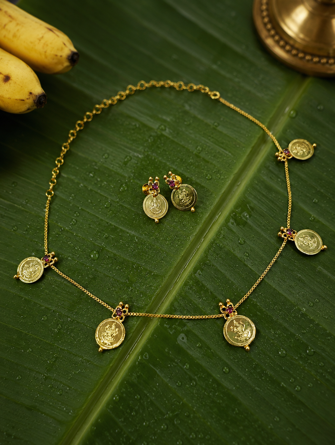 Traditional South Indian gold-plated Mookambika Kasu Mala coin necklace and matching stud earrings with ruby accents, displayed on a fresh green banana leaf with water droplets.