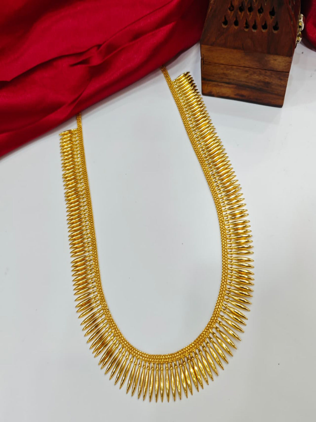 Traditional South Indian gold long haram necklace featuring an intricate Mallige Moggu or Mullu Mottu jasmine bud design, styled on a white surface next to a red silk fabric and a small wooden box.