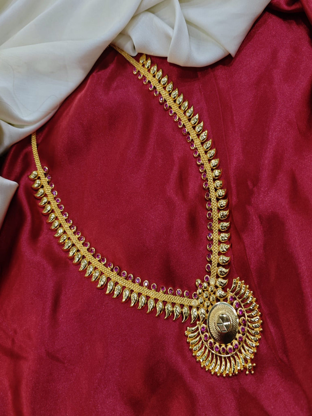 South Indian traditional gold Maanga Mala long haram necklace featuring a mango leaf chain motif with ruby pink stones and a large round textured pendant, displayed on red and white silk fabric.