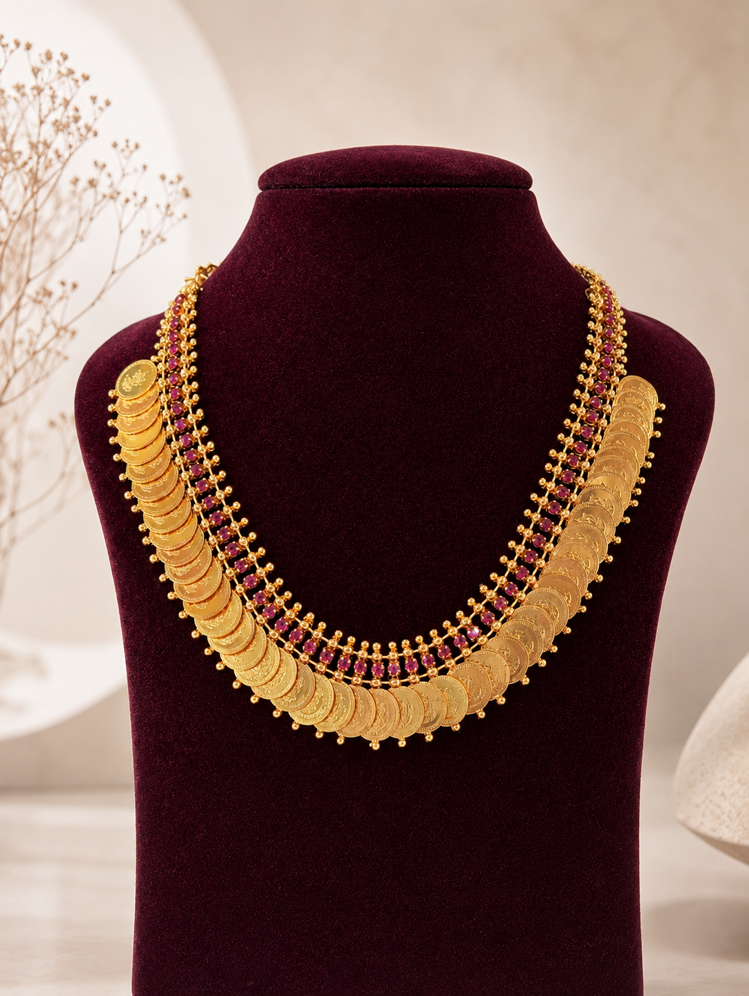 Traditional South Indian gold coin necklace known as Kasu Mala or Kasulaperu named Vishalaakshi, featuring rows of overlapping engraved gold coins with a multi-layered border of small pink rubies, displayed on a deep burgundy velvet jewelry bust.