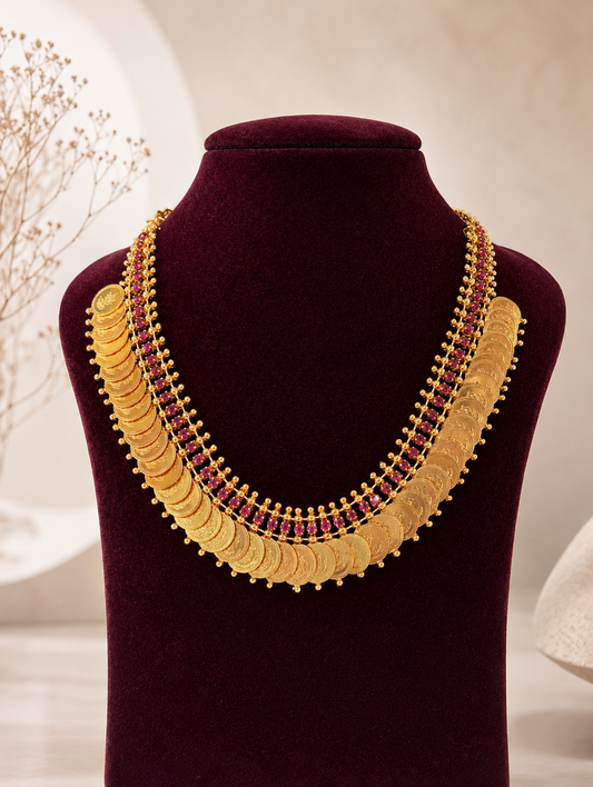 Traditional South Indian gold coin necklace known as Kasu Mala or Kasulaperu named Vishalaakshi, featuring rows of overlapping engraved gold coins with a multi-layered border of small pink rubies, displayed on a deep burgundy velvet jewelry bust.