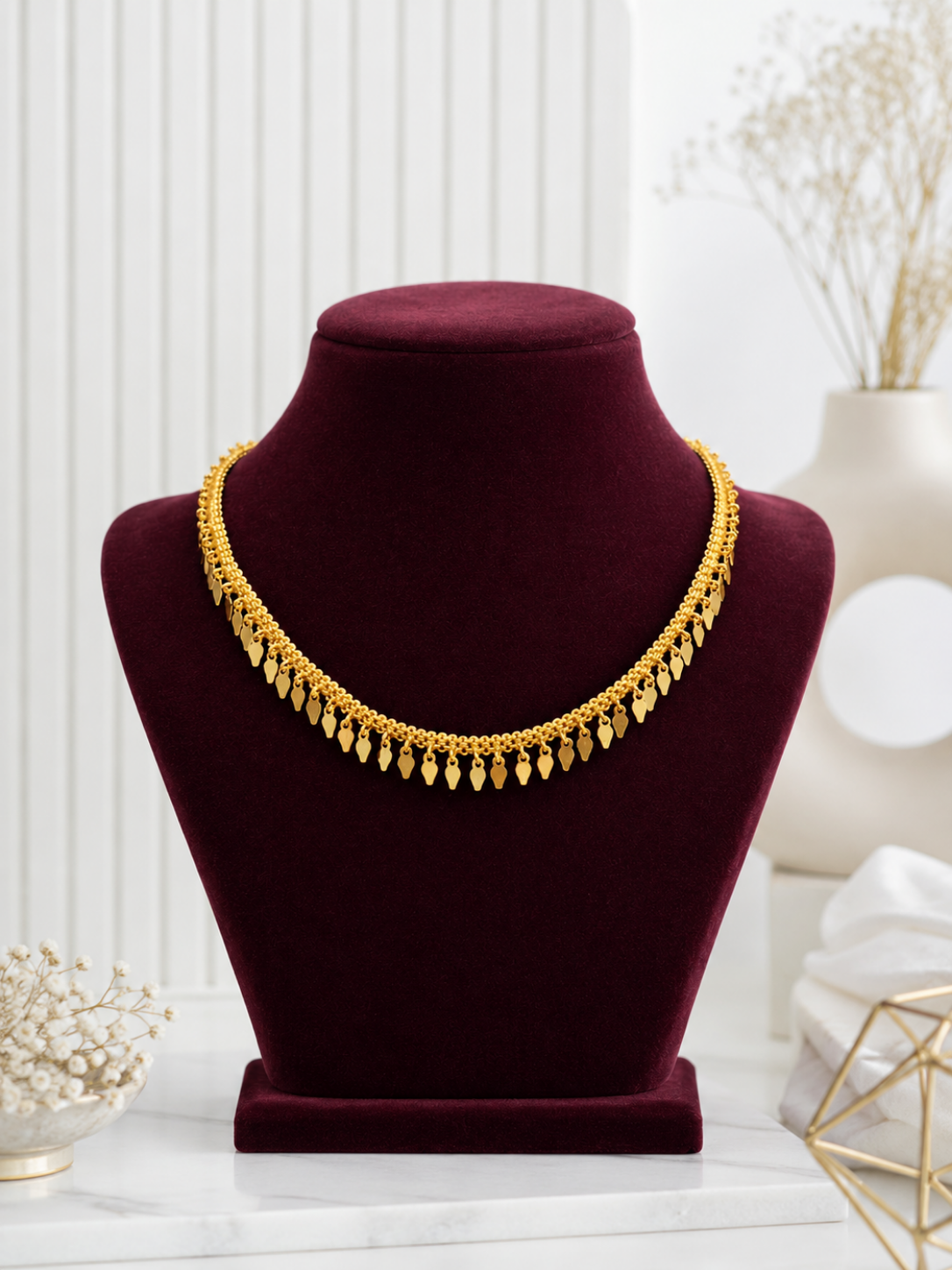 A minimalist gold choker necklace named Saroja, featuring a textured chain accented with a row of small, dangling leaf-shaped gold charms, elegantly displayed on a deep burgundy velvet jewelry bust.