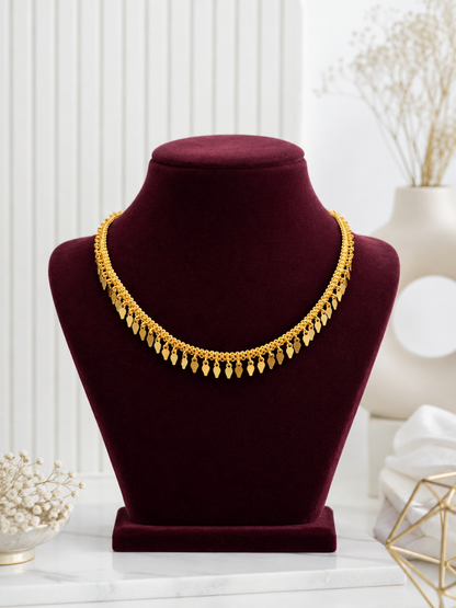 A minimalist gold choker necklace named Saroja, featuring a textured chain accented with a row of small, dangling leaf-shaped gold charms, elegantly displayed on a deep burgundy velvet jewelry bust.